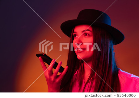 One young beautiful girl wearing black hat using phone isolated on dark studio background in neon light. Concept of emotions, fashion 83540000
