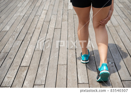 Rear view of unrecognizble female runner in blue sports shoes standing on wooden pier and touching pain point while feeling pain under kneecap 83540307