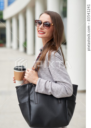 Portrait of cheerful city girl with black leather bag drinking takeout coffee on move outdoors Portrait of cheerful city girl with black leather bag drinking takeout coffee on move outdoors 83541241