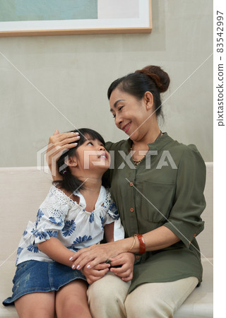 Smiling loving Asian grandma with hair bun sitting on sofa and stroking granddaughter while cuddling her Smiling loving Asian grandma with hair bun sitting on sofa and stroking granddaughter while cuddling her 83542997