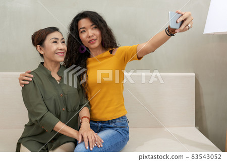 Attractive curly-haired young Vietnamese woman sitting on sofa and taking selfie with mature mother on smartphone 83543052