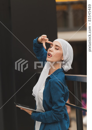 Portrait of an Arab student girl holding a tablet. Arab business woman in hijab holding a tablet in the street. Woman is dressed in hijab Portrait of an Arab student girl holding a tablet. Arab business woman in hijab holding a tablet in the street. Woman is dressed in hijab 83545489
