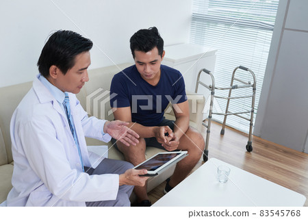 Horizontal high angle shot of mature Asian doctor wearing white coat showing something to his young male patient on digital tablet 83545768