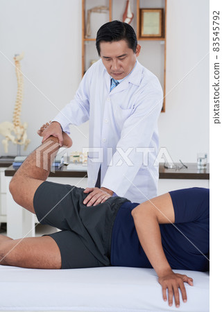 Vertical medium long shot of mature Asian physical therapist wearing white coat working with unrecognizable male patient recovering his hip joints range of motion 83545792