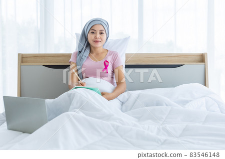 a asian women disease mammary cancer patient with pink ribbon wearing headscarf After treatment to chemotherapy with Take notes in a notebook and laptop on bed In the bedroom at the house,healthcare 83546148