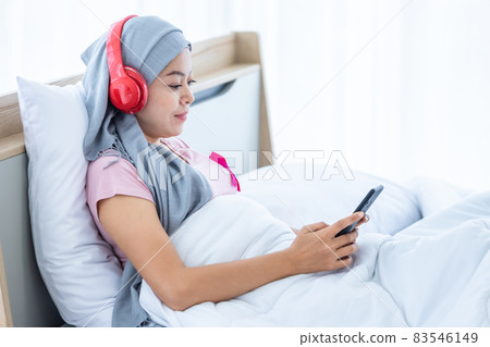 Happy a asian women disease mammary cancer patient with pink ribbon wearing headscarf in headphones is listening to music with smartphone After treatment to chemotherapy sit on bed In the bedroom 83546149