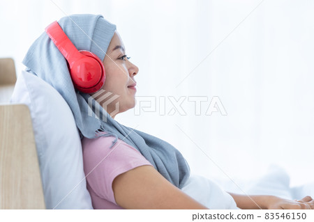 Happy a asian women disease mammary cancer patient with pink ribbon wearing headscarf in headphones is listening to music After treatment to chemotherapy sit on bed In the bedroom at the house Happy a asian women disease mammary cancer patient with pink ribbon wearing headscarf in headphones is listening to music After treatment to chemotherapy sit on bed In the bedroom at the house 83546150
