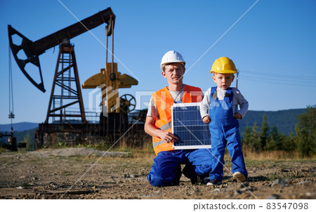 Engineer in waistcoat and helmet keeping solar panel in hands next to kid boy. Experienced mechanic and his little follower against of oil rig. Solar energy is ecological alternative to oil industry. Engineer in waistcoat and helmet keeping solar panel in hands next to kid boy. Experienced mechanic and his little follower against of oil rig. Solar energy is ecological alternative to oil industry. 83547098