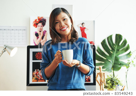 Portrait of positive young Asian magazine designer in blue sweater holding mug against computer and banners in office 83549243