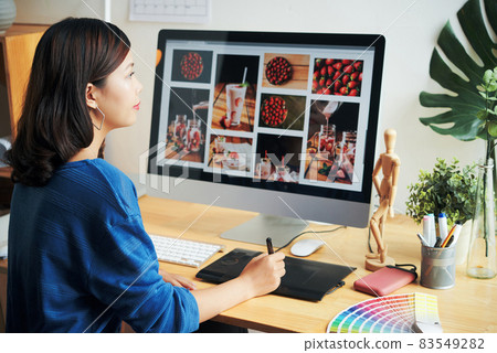 Close-up of busy young Asian designer sitting at wooden desk with color swatch and using digitizer while retouching photos 83549282