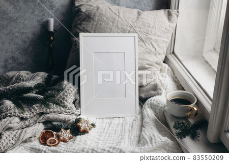 Christmas still life. Candle in golden candleholder. White picture frame mockup. linen pillow near window. Moody composition. Fir tree branches, cup of coffee and gingerbread cookies. Winter interior. 83550209