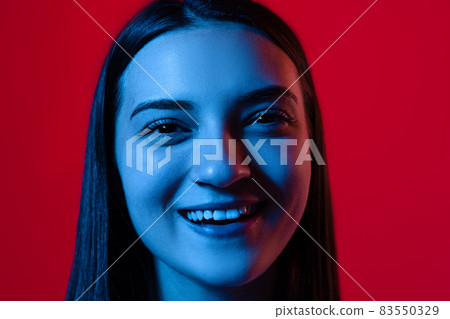 Close-up portrait of young beautiful woman smiling, posing isolated over red background in neon lights 83550329