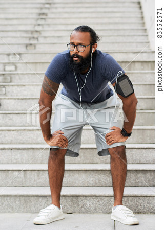 Exhausted young Indian man with beard leaning hands on knees while resting after running on stairs Exhausted young Indian man with beard leaning hands on knees while resting after running on stairs 83552571
