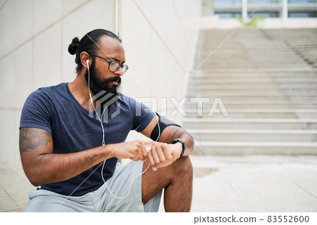 Serious young bearded Indian man with tattoo sitting at corner of building and checking fitness watch after training Serious young bearded Indian man with tattoo sitting at corner of building and checking fitness watch after training 83552600