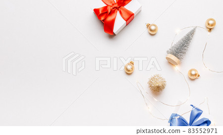 Holiday winter background. White gift box with red ribbon, New Year balls and sparkling lights garland in Christmas composition on white for greeting card. Flat lay, top view, copy space. 83552971