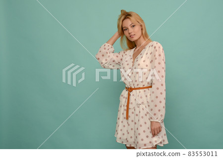 Delighted beautiful young woman in white dress in polka dots posing in studio on blue background. 83553011