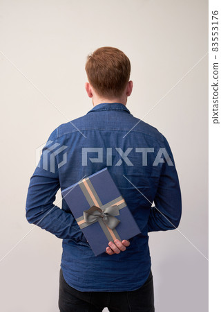 Rear view of redhead man in shirt hiding present box with bow tie behind back, isolated background 83553176