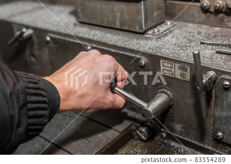The hand of a man in a black work jumpsuit controls a milling machine by the handle for processing metal in an industrial enterprise The hand of a man in a black work jumpsuit controls a milling machine by the handle for processing metal in an industrial enterprise 83554289