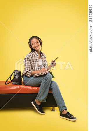 cute girl laughing at camera wearing headphones and writing using pen in book while holding cell phone 83556922