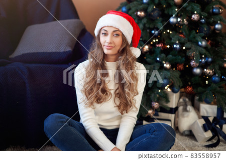 Christmas. Woman dressed white sweater and jeans sitting on the floor near christmas tree with present box Christmas. Woman dressed white sweater and jeans sitting on the floor near christmas tree with present box 83558957