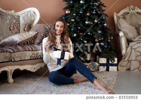 Christmas. Woman dressed white sweater and jeans sitting on the floor near christmas tree with present box Christmas. Woman dressed white sweater and jeans sitting on the floor near christmas tree with present box 83558958