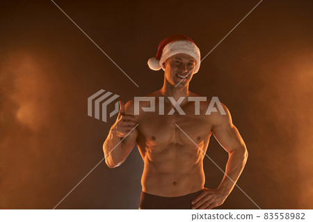 Portrait of muscular man wearing Christmas Santa hat, showing thumb up on smoky background 83558982