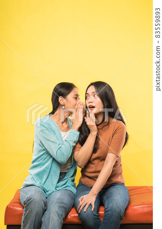 a beautiful woman is surprised when her friend whispers while chatting sitting together on the couch 83559893