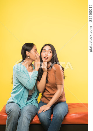 a young asian woman with a surprised gesture as her friend whispers while sitting together 83560013
