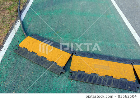 Black and yellow heavy plastic covers for people and vehicle to cross over cables laid on road. Pavement pads to protect electrical wiring on sidewalk in public park. Channel for laying the wires. Black and yellow heavy plastic covers for people and vehicle to cross over cables laid on road. Pavement pads to protect electrical wiring on sidewalk in public park. Channel for laying the wires. 83562504