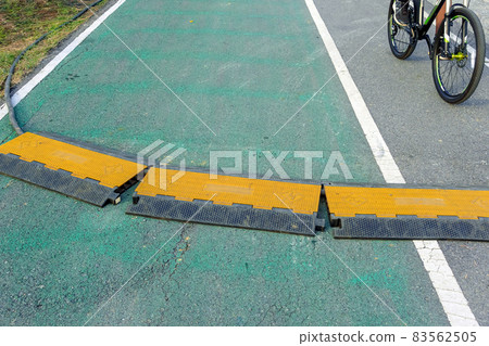 Black and yellow heavy plastic covers for people and vehicle to cross over cables laid on road. Pavement pads to protect electrical wiring on sidewalk in public park. Channel for laying the wires. 83562505