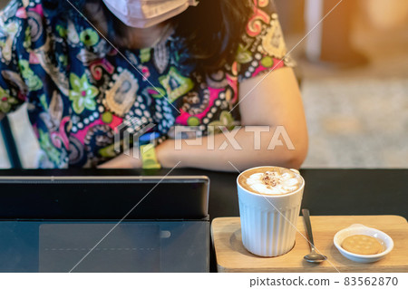 Hot Cappuccino coffee for quarantined woman wear face mask and work from home by using tablet and stress relief with hot coffee. Concepts of Covid-19 (Coronavirus disease). Selective focus on coffee. 83562870