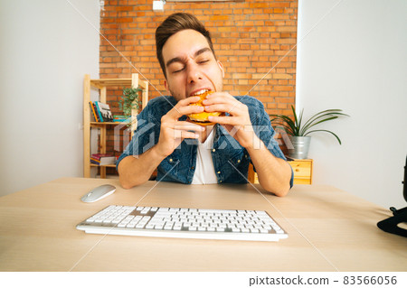 Front view of handsome hungry young man eating hamburger with beef from fast food restaurant sitting at desk with wireless keyboard 83566056