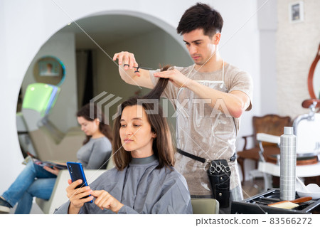 Girl client using phone during hair cut by professional hairdresser Girl client using phone during hair cut by professional hairdresser 83566272