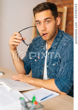 Vertical shot of focused serious young man reading paper book holding end of glasses near mouth sitting at desk in home office, looking at camera. 83566343
