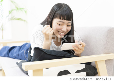 一位年輕女子躺在家裡的沙發上,用智能手機看在線電視 一位年輕女子躺在家裡的沙發上,用智能手機看在線電視 83567125
