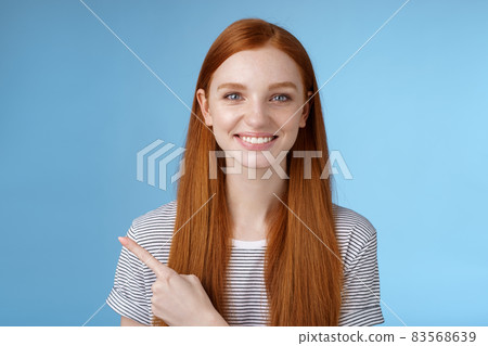 Pleasant charismatic helpful friendly-looking female redhead blue eyes pointing left index finger help make decision pick product store smiling broadly confident gaze camera asking trust her 83568639
