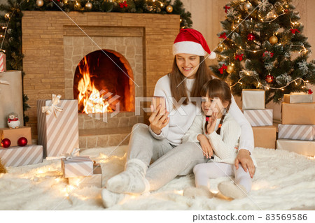 Mother and daughter using smart phone for video call, talking with someone, congratulating with X'Mas, posing in room with Christmas tree and fireplace, family sitting on floor on soft carpet. Mother and daughter using smart phone for video call, talking with someone, congratulating with X'Mas, posing in room with Christmas tree and fireplace, family sitting on floor on soft carpet. 83569586