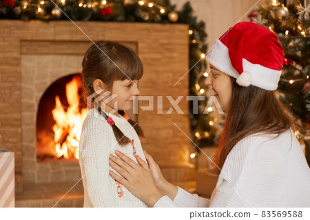 Happy family mother and little daughter playing in Christmas eve, posing near fireplace, mom telling her little girl something interesting, wearing white sweaters. Happy family mother and little daughter playing in Christmas eve, posing near fireplace, mom telling her little girl something interesting, wearing white sweaters. 83569588