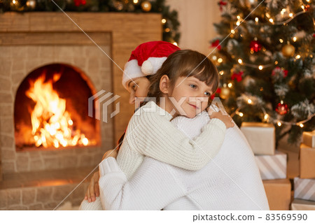 Mother and child hugs on winter evening by fireplace, dreamily female kid wearing white sweater being satisfied spend Christmas Eve with mommy, posing in living room near xmas tree. Mother and child hugs on winter evening by fireplace, dreamily female kid wearing white sweater being satisfied spend Christmas Eve with mommy, posing in living room near xmas tree. 83569590