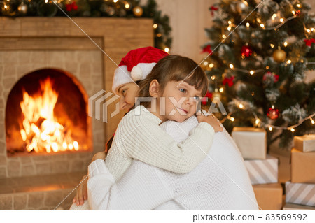 Happy mommy and little charming daughter hugging in festive living room near Christmas tree and fireplace, family celebrating New Year Eve at home. Happy mommy and little charming daughter hugging in festive living room near Christmas tree and fireplace, family celebrating New Year Eve at home. 83569592