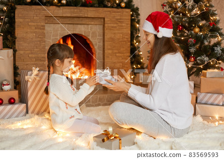 Mother giving present box to her little daughter, smiling girl looks with satisfied expression on her gift, ladies wearing white shirts, sitting near fireplace and fir tree. Mother giving present box to her little daughter, smiling girl looks with satisfied expression on her gift, ladies wearing white shirts, sitting near fireplace and fir tree. 83569593