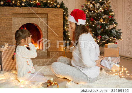 Young caucasian mother and her cute daughter giving christmas gift for her he little daughter, child covering eyes with palms, family wearing white sweaters. Young caucasian mother and her cute daughter giving christmas gift for her he little daughter, child covering eyes with palms, family wearing white sweaters. 83569595