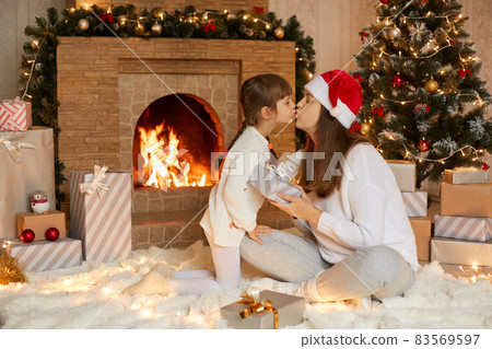 Happy mother and daughter giving christmas gifts, mom kisses her little girl while sitting on floor with crossed legs, female with child posing in festive living room. Happy mother and daughter giving christmas gifts, mom kisses her little girl while sitting on floor with crossed legs, female with child posing in festive living room. 83569597