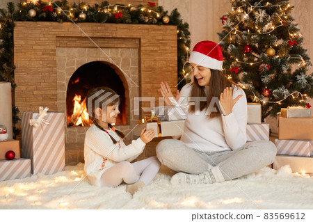 Charming female kid presenting Christmas gift to her mother at Christmas morning, celebrating Christmas and New Year, happy time together, surprised mom being glad to get cool present. 83569612