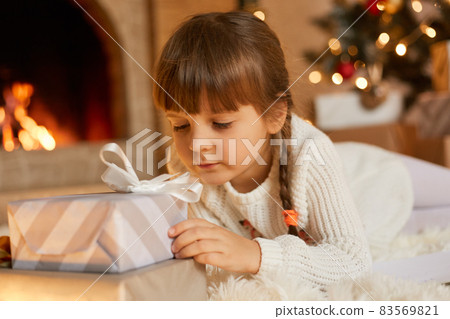 Pensive child with christmas present at home, female kid looks at gift box with ribbon and dreaming to open it, little girl with pigtails posing in living room near xmas tree and fireplace. Pensive child with christmas present at home, female kid looks at gift box with ribbon and dreaming to open it, little girl with pigtails posing in living room near xmas tree and fireplace. 83569821