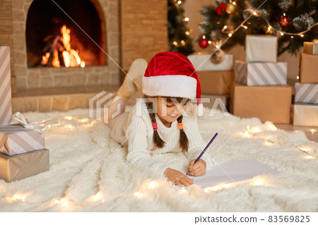 Little cute female child lying on floor on carpet and writing letter to Santa Claus, looking at empty paper and think, posing against present boxes, xmas tree and fire places. Little cute female child lying on floor on carpet and writing letter to Santa Claus, looking at empty paper and think, posing against present boxes, xmas tree and fire places. 83569825