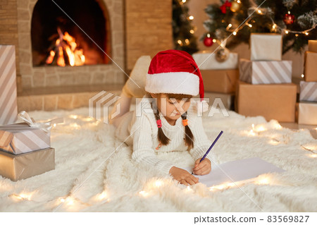 Little girl in red christmas hat writes letter to Santa Claus, adorable child lying on floor near fireplace, wearing white warm jumper, lying on floor on soft carpet. Little girl in red christmas hat writes letter to Santa Claus, adorable child lying on floor near fireplace, wearing white warm jumper, lying on floor on soft carpet. 83569827