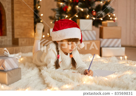 Cute child girl writing letter santa at home near gift boxes while lying on floor on soft carpet, kid with pigtails drawing picture for present, poses near xmas tree. Cute child girl writing letter santa at home near gift boxes while lying on floor on soft carpet, kid with pigtails drawing picture for present, poses near xmas tree. 83569831
