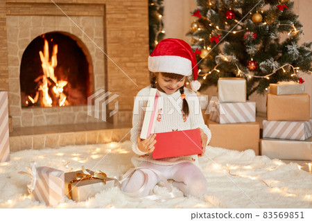 Happy small girl opening Christmas present box, looks inside with smile, being happy to get such gift, wearing white pullover and santa claus hat, sitting on floor in festive room with xmas decoration 83569851
