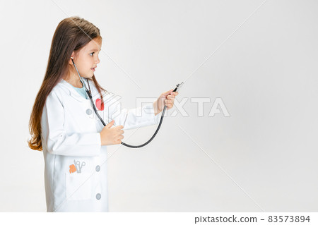 Close-up portrait of cute little girl, child in image of doctor wearing white lab coat with stethoscope isolated on white studio background Close-up portrait of cute little girl, child in image of doctor wearing white lab coat with stethoscope isolated on white studio background 83573894